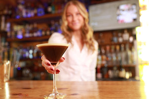 A female bartender holding out in front of her a non-alcoholic espresso martini cocktail made from Nitro Mocktails Espress0% Martini in a bar setting