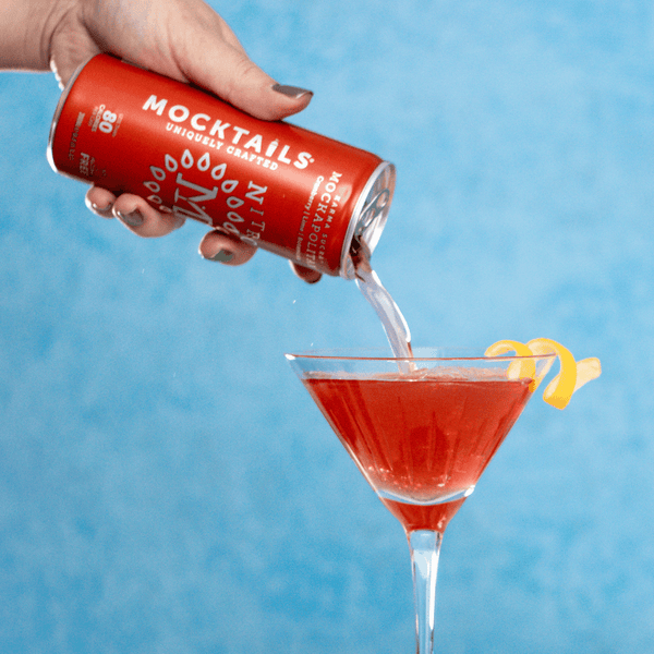 Nitro Mocktails Mockapolitan mocktail being poured into a martini glass with lemon twist garnish on blue background.