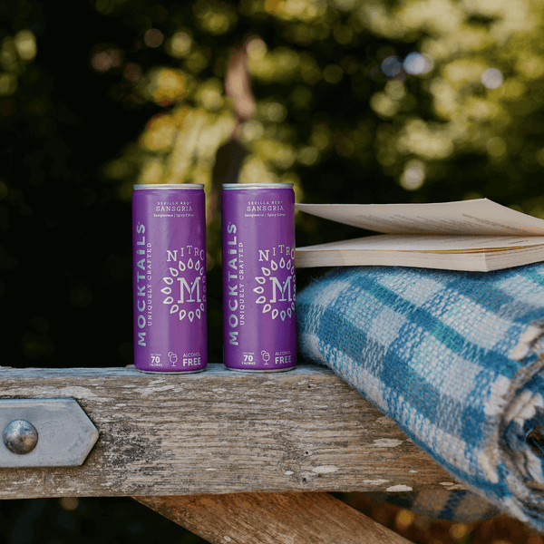 Nitro Mocktails Sansgria non-alcoholic cocktail cans on outdoor bench with plaid blanket and book—alcohol-free sangria for relaxed, mindful moments.