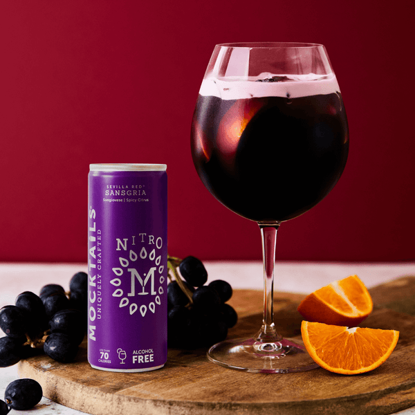 Sansgria non-alcoholic sangria cocktail in wine glass beside Nitro Mocktails can, with fresh grapes and orange slices on a wooden board.