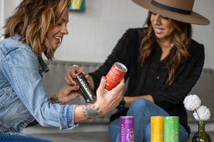 Two women holding and observing non-alcoholic Nitro Mocktails cans in a cafe setting