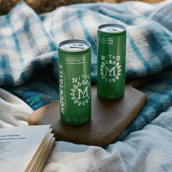 Two cans of Nitro Mocktails Mockarita on a wooden tray with a cozy blanket—vegan-friendly, alcohol-free margarita mocktail outdoors.