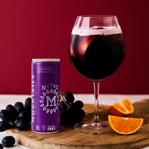 Sansgria non-alcoholic sangria cocktail in wine glass beside Nitro Mocktails can, with fresh grapes and orange slices on a wooden board.